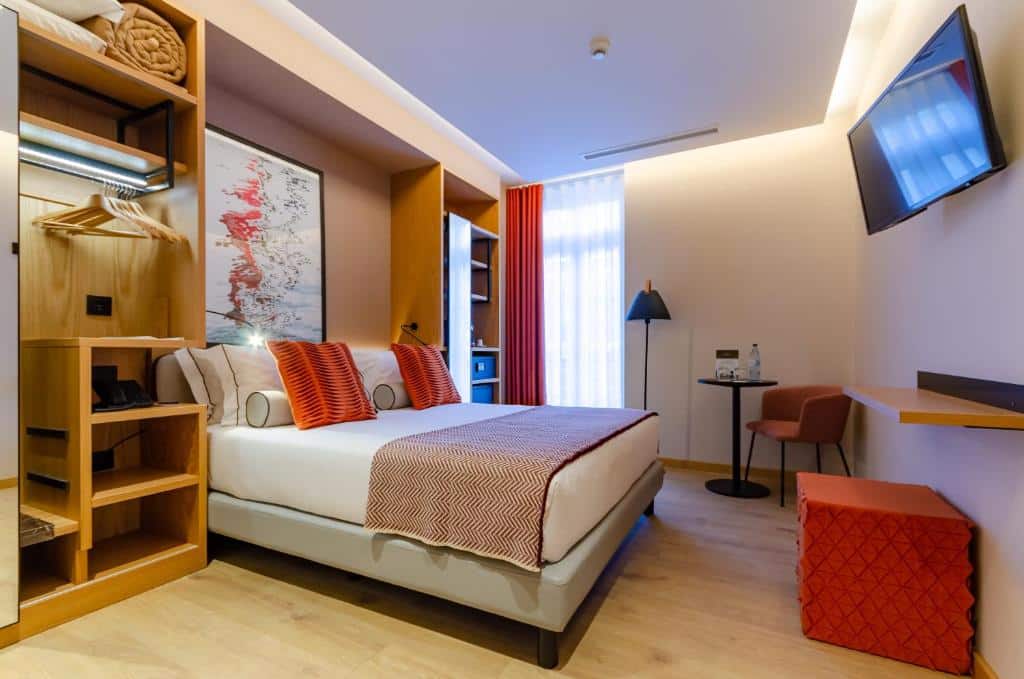 Quarto do Rossio Plaza Hotel com cama de casal, varanda com cortinas, cadeirão estofado junto a uma pequena mesa e candeeiro de pé; em redor da cama, um móvel com cabide, prateleiras e espelho, e em frente à cama uma televisão