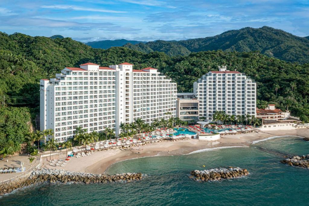 Vista aérea de dois grandes edifícios do Hilton Vallarta Riviera à beira-mar com piscinas, palmeiras e uma praia rodeada por montanhas verdes.