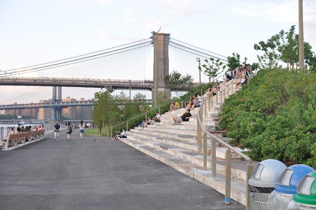 Amplo caminho para peões no Brooklyn Bridge Park, com degraus onde várias pessoas estão sentadas e uma ponte suspensa ao fundo.