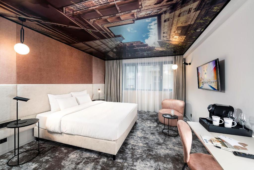 Quarto do Three Corners Downtown Hotel com uma cama grande, cadeiras cor-de-rosa, secretária, TV e um mural no teto com um edifício e céu azul.