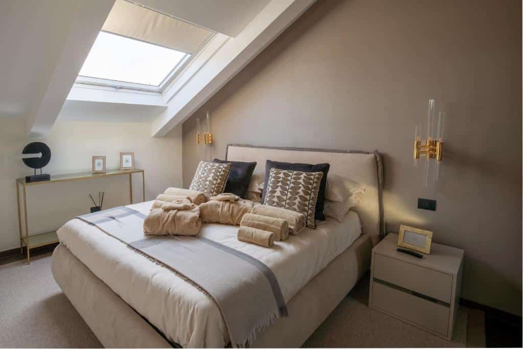 Quarto amplo do Margherita Apartments com cama de casal, cabeceira estofada, mesas de cabeceira, candeeiro de parede dourado e decoração em tons neutros.