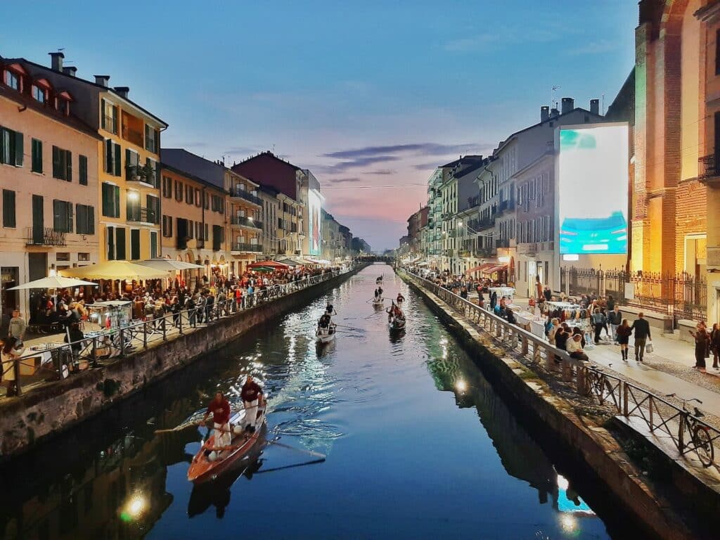 Um dos canais de Navigli, em Milão, com barcos a passar e, nas margens, comércio e pessoas a passear