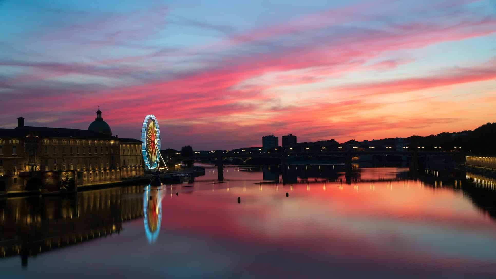 Roda-gigante e edifícios junto a um rio ao pôr do sol, com o céu rosa e roxo refletido na água, em Toulouse.