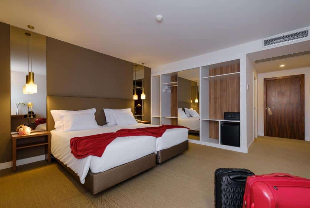 Quarto moderno no Hotel Regina com duas camas individuais, mantas vermelhas, mesas de cabeceira, roupeiro aberto e duas malas no chão.