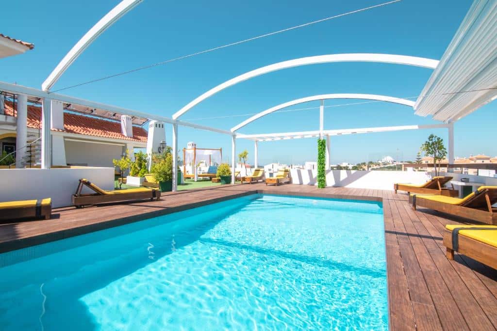 Piscina exterior retangular com água azul-clara, rodeada de espreguiçadeiras e deck de madeira sob estruturas brancas em arco, no Loulé Jardim Hotel em Loulé.