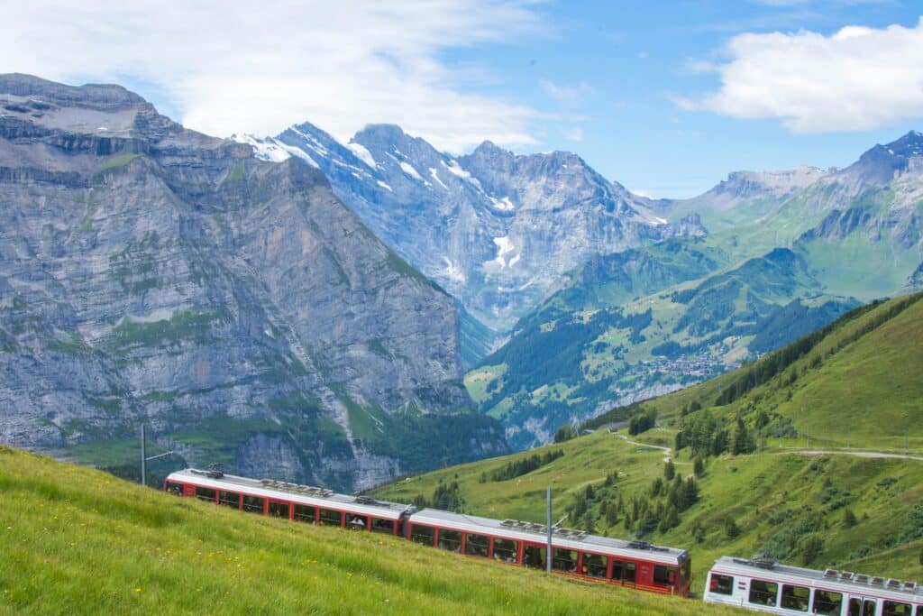 Um comboio vermelho percorre uma paisagem montanhosa verdejante com picos rochosos ao fundo, sob um céu azul. Representa como viajar de comboio pela Suíça.