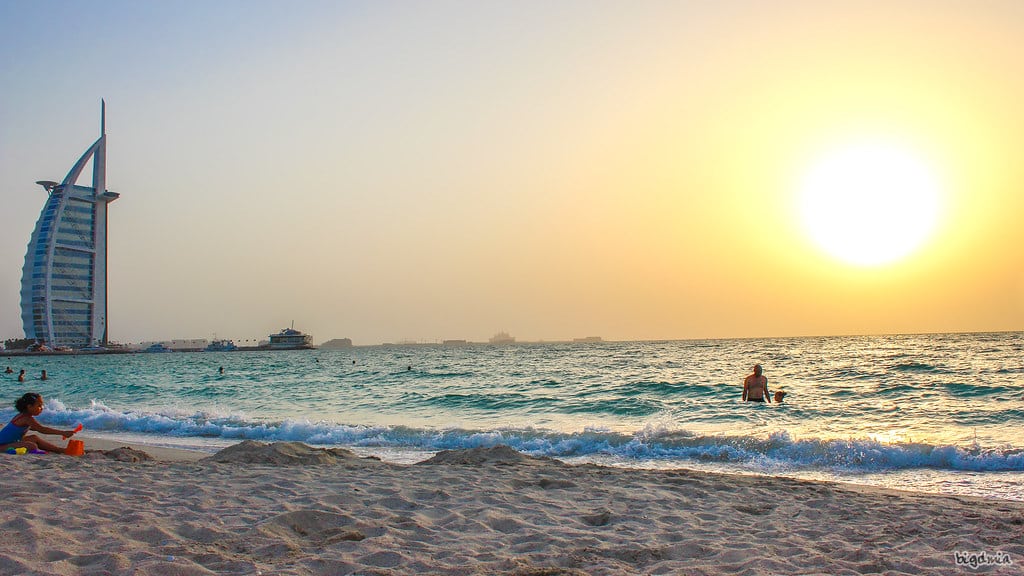 A Jumeirah Public Beach ao pôr do sol, com o mar à vista, pessoas a nadar e um edifício alto em forma de vela.