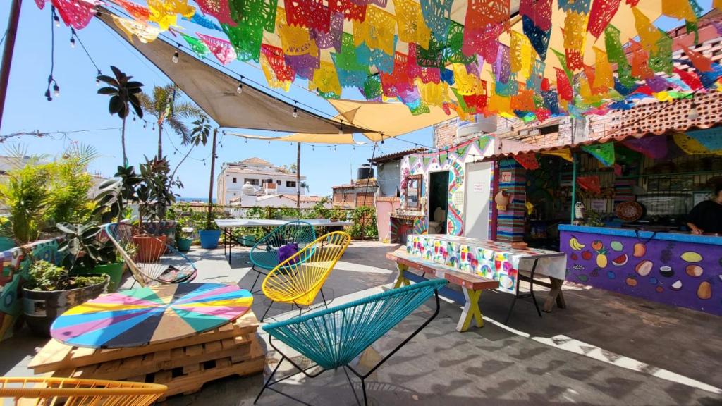 Pátio colorido ao ar livre do Hostel Vallarta com bandeirolas festivas de papel picado, cadeiras vibrantes, mesas e plantas sob chapéus de sol.