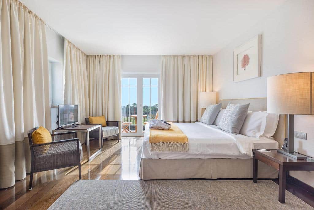 Quarto no The Lake Resort em Vilamoura, bem iluminado, com cama de casal, poltrona, secretária, TV, janela grande com cortinas e decoração neutra.