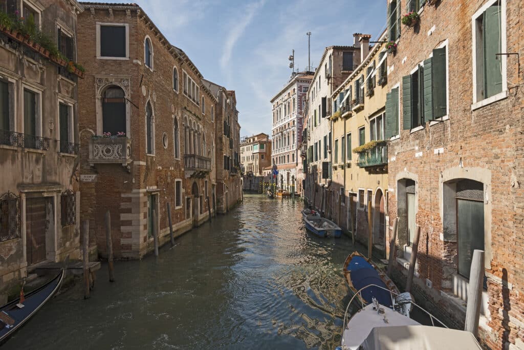 Ruas estreitas de Veneza atravessadas por canais e pontes, com edifícios residenciais de tijolo, janelas com portadas verdes e barcos típicos a flutuar na água
