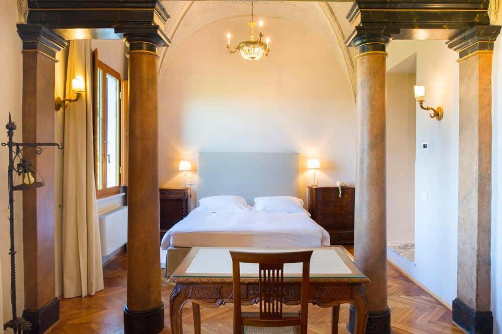 Quarto de hotel clássico com cama de casal, colunas e lustre em ambiente iluminado