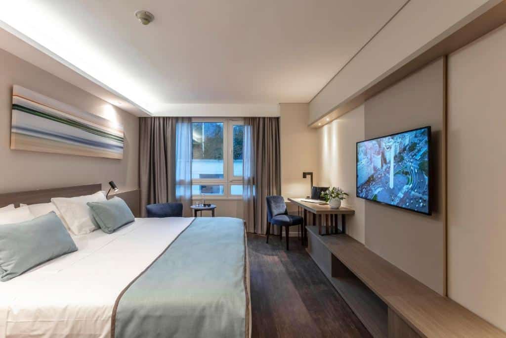 quarto do Huinid Obelisco Hotel com cama de casal grande, tv em frente, mesas de cabeceira dos dois lados e janela ampla com cortinas