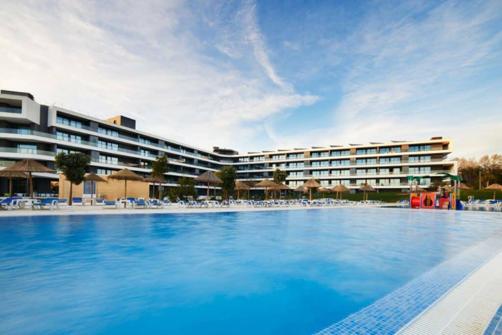 Ampla piscina externa em frente ao edifício do RR Alvor Baía Resort de vários andares do hotel com espreguiçadeiras e guarda-sóis.