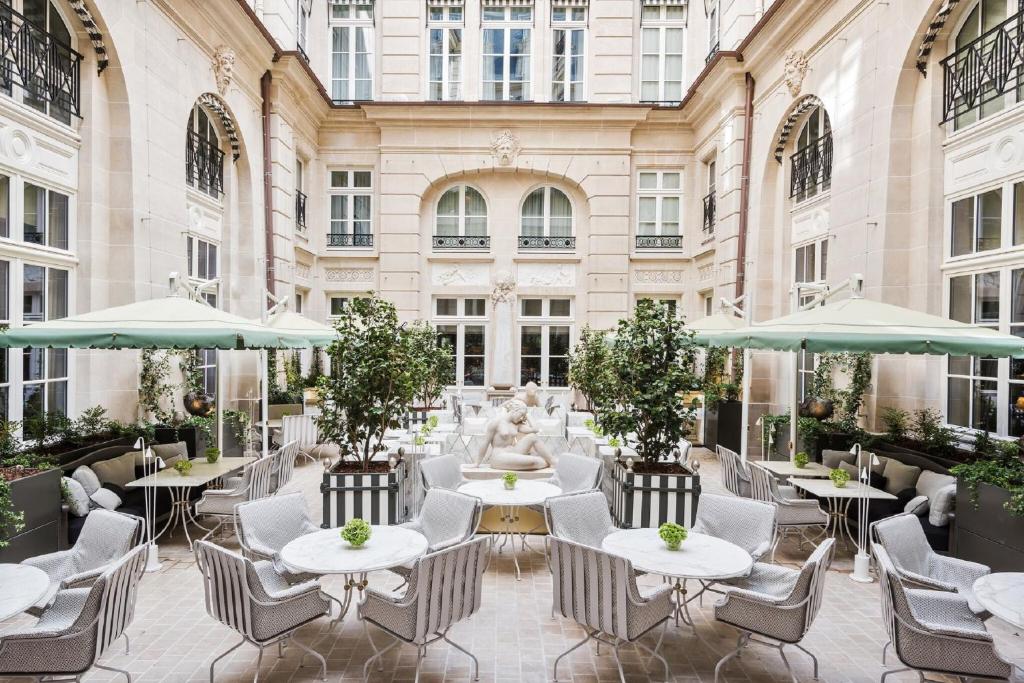 Pátio exterior elegante do Hotel de Crillon com mesas brancas, cadeiras, chapéus de sol verdes, plantas e uma estátua central, rodeado por edifícios altos. Este é um dos hotéis de luxo em Paris.