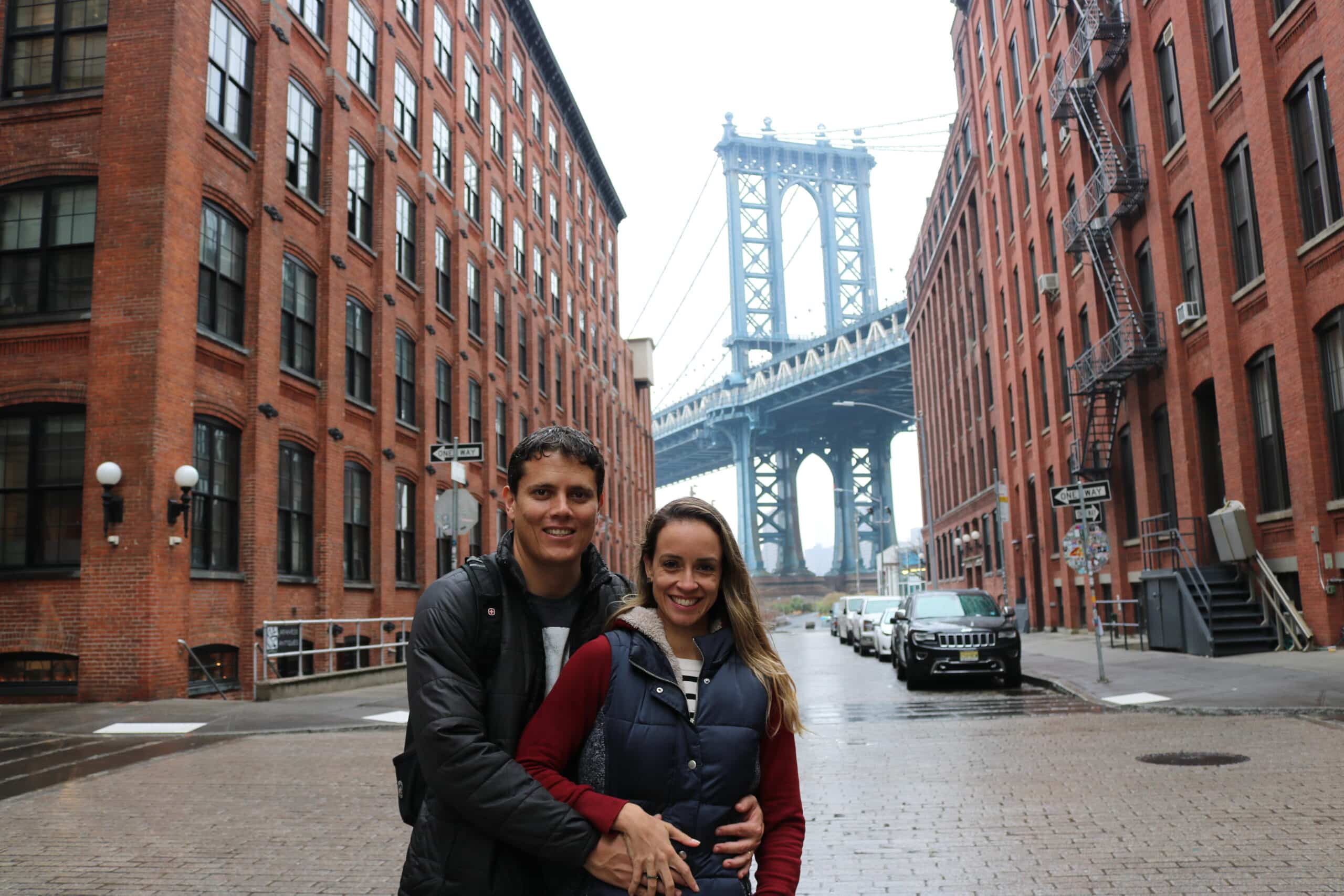 Duas pessoas de pé no meio de uma rua de Dumbo, ladeada por prédios, com a Ponte de Manhattan ao fundo.