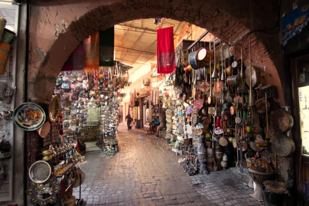Um beco estreito das ruas da Medina de Marraquexe, com vários produtos pendurados, artigos de metal e cerâmica, sob uma entrada em arco que leva para dentro do bazar.