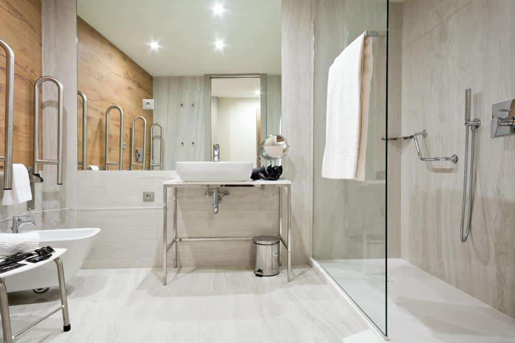 Casa de banho adaptada do Acta The Avenue com pavimento antiderrapante, lavatório baixo e sanita com barras de apoio e, do lado direito, chuveiro com barra de apoio.