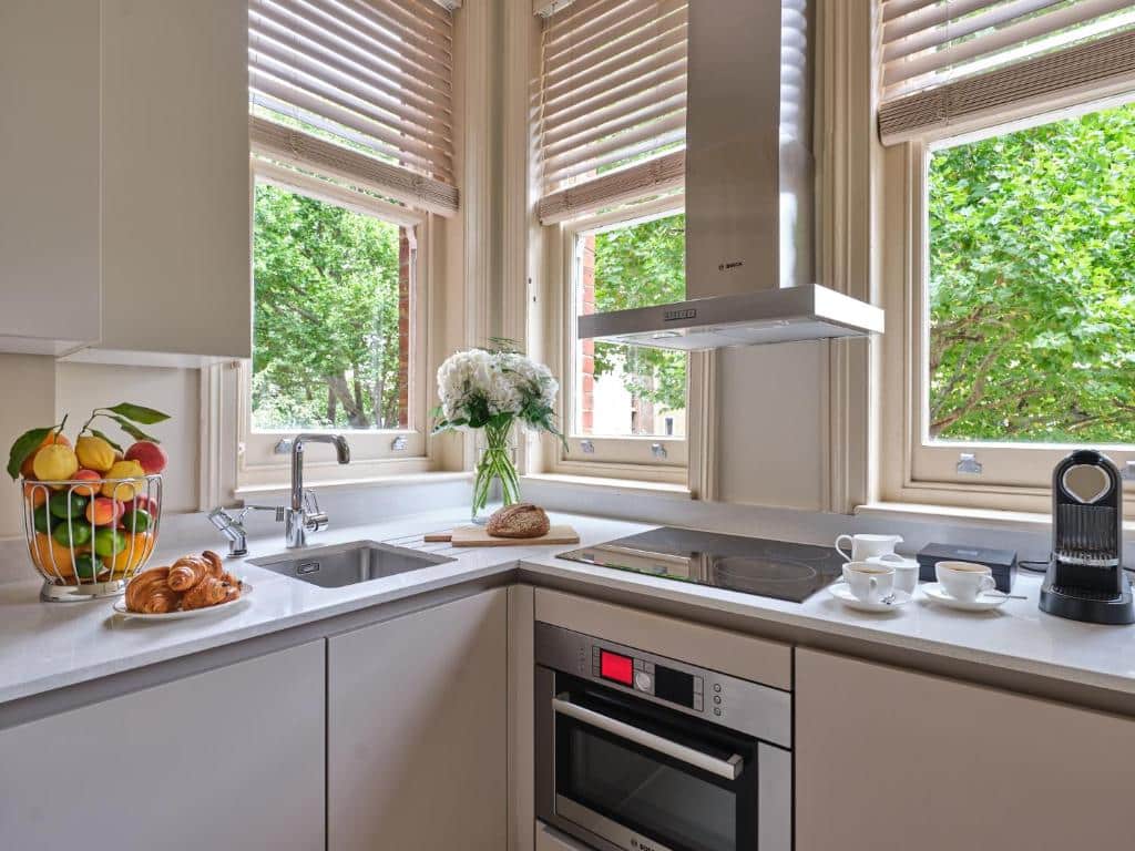 Cozinha do The Apartments by The Sloane Club com três janelas com estores, alguns vasos de flores, uma máquina de café, um forno e um cesto com fruta.
