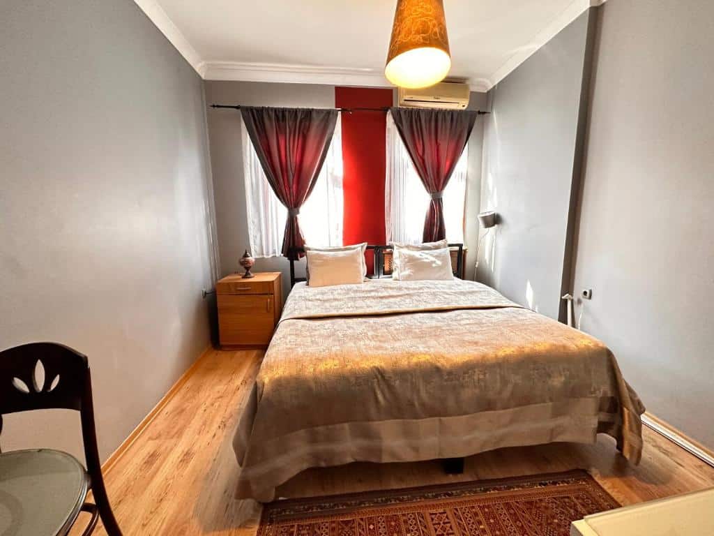 Uma cama de casal bem arrumada num quarto pequeno do Ahmet Efendi Evi, com paredes cinzentas, pavimento de madeira, duas janelas e um candeeiro suspenso.