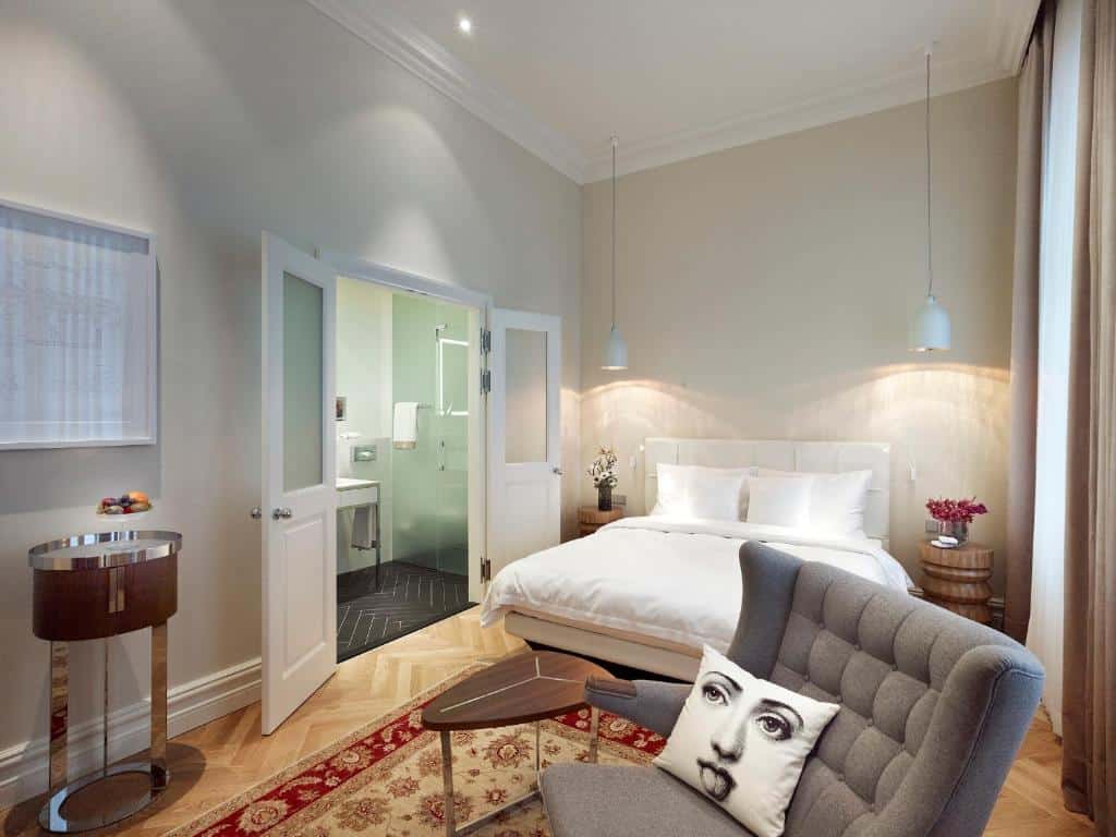 Quarto moderno no Hotel Sans Souci Wien, com cama de casal, poltrona, tapete estampado e casa de banho privativa com portas de vidro.