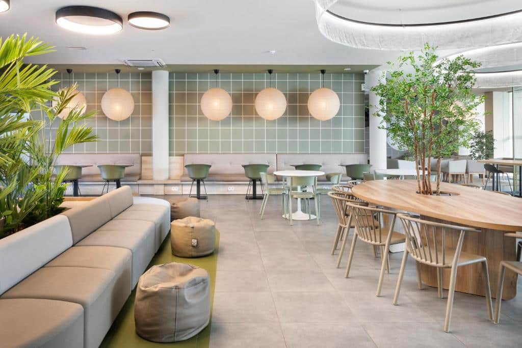 Zona comum do B&B HOTEL Lisboa Aeroporto, com várias mesas e cadeiras, um sofá do lado esquerdo, bufetes e alguns vasos de plantas.