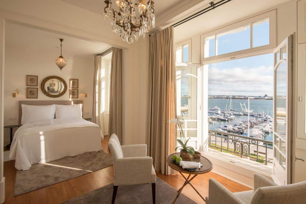 Quarto elegante no Grand House – Relais & Chateaux, em Vila Real de Santo António, com cama, cadeiras, lustre e janelas grandes com vista para a marina e a água.
