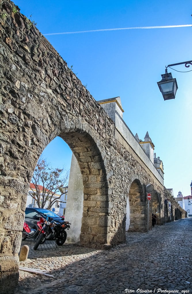 Arcos antigos de pedra em rua de calçada com motociclos e casas coloridas em cidade histórica portuguesa. Ilustra este artigo sobre o que fazer em Évora.