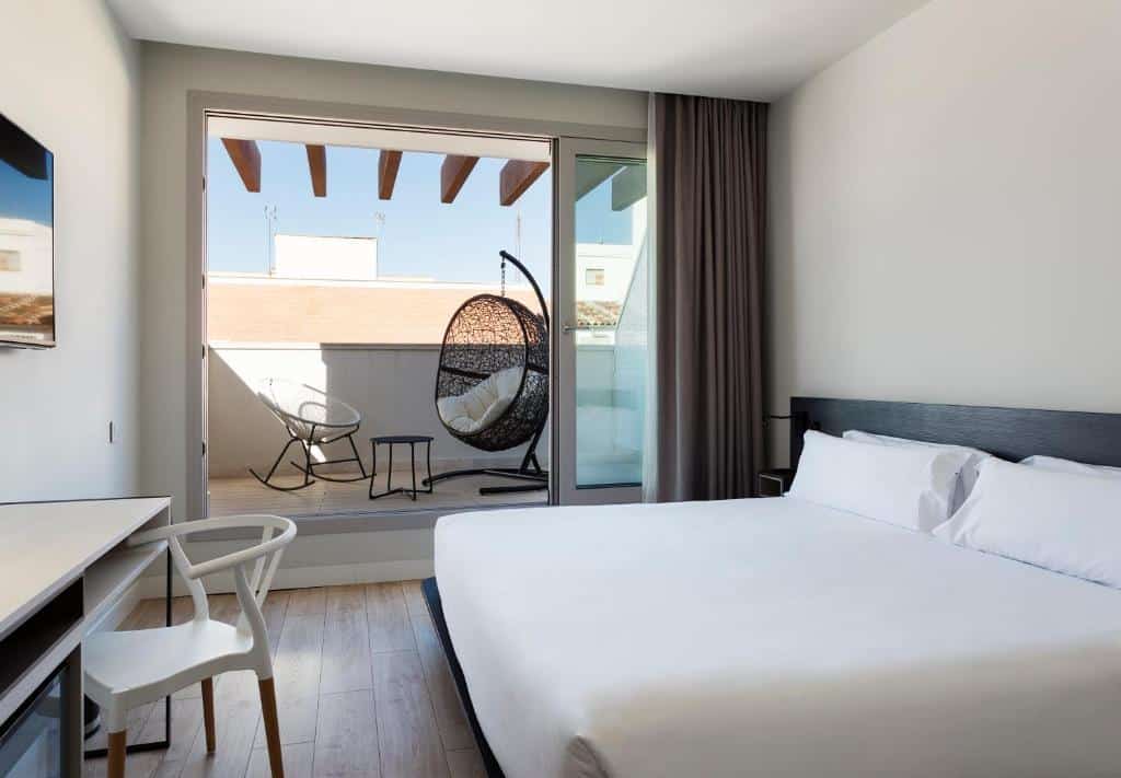 Quarto do B&B Hotel Madrid Centro Puerta del Sol, um dos melhores hotéis em Madrid. A cama de casal está encostada à parede do lado esquerdo e fica em frente a uma secretária, com cadeira e uma TV na parede por cima. Ao fundo, uma grande porta de vidro com cortinas no canto está aberta e dá acesso a um terraço com poltronas.