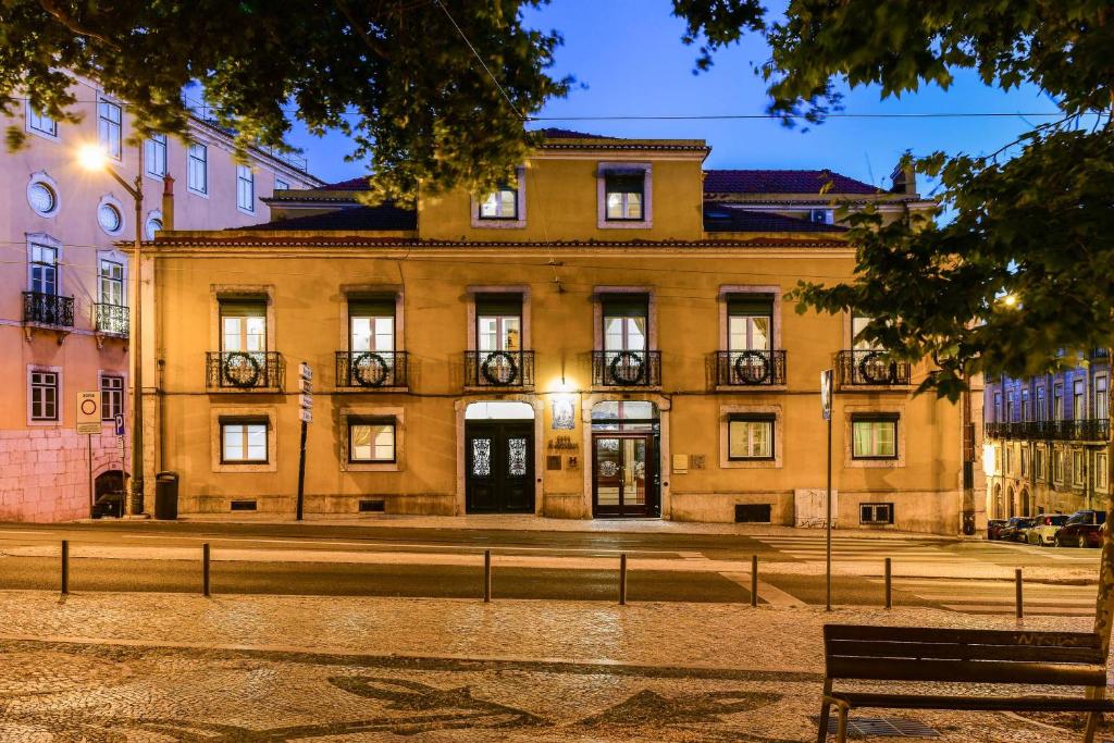 Edifício amarelo de dois pisos, onde fica a Casa de São Mamede Hotel, com janelas iluminadas ao anoitecer, visto de uma rua de calçada com árvores.