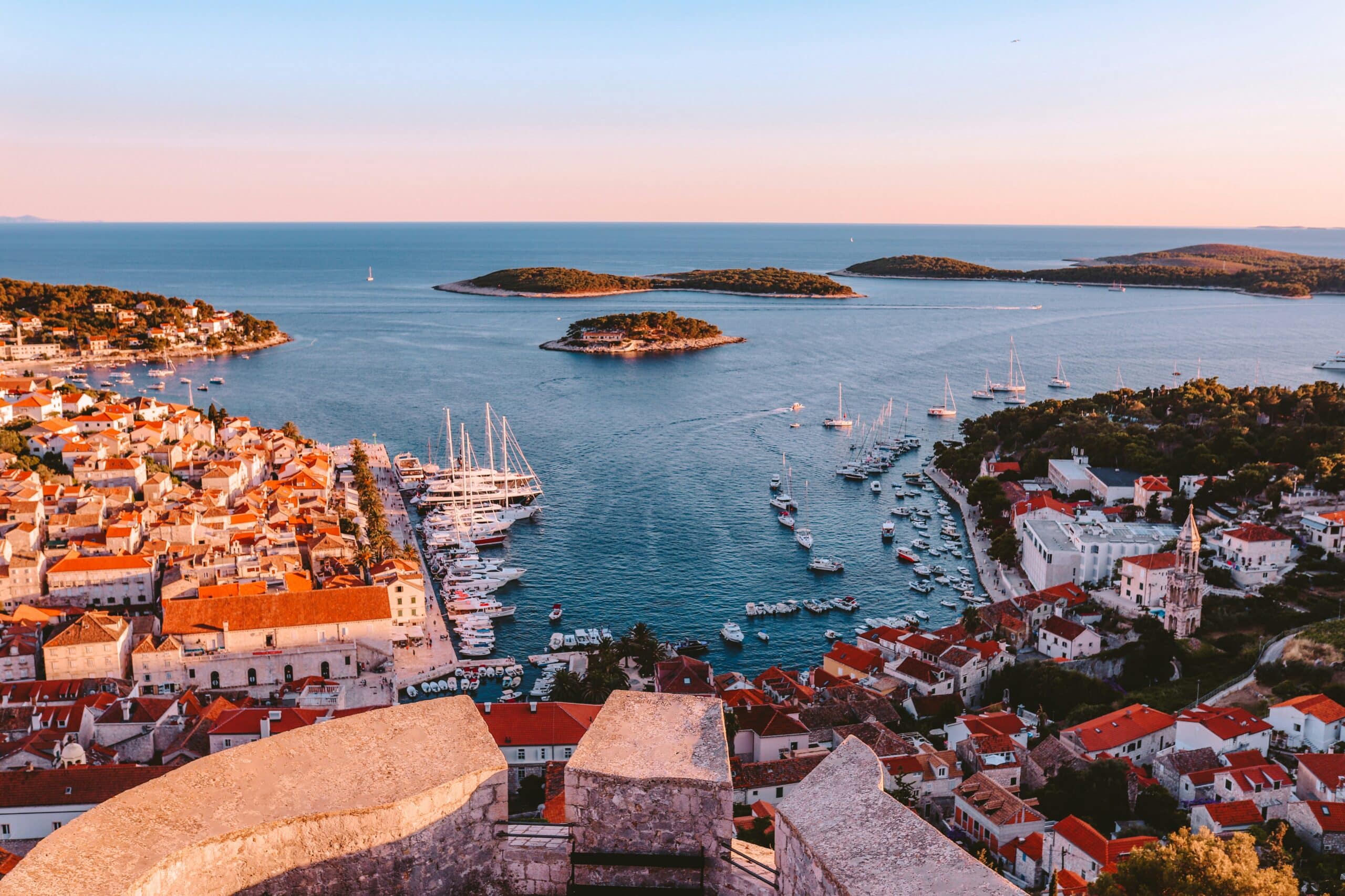 Vista aérea da cidade costeira de Hvar, com edifícios de telhados vermelhos, marinas e várias pequenas ilhas num mar azul calmo ao pôr do sol.