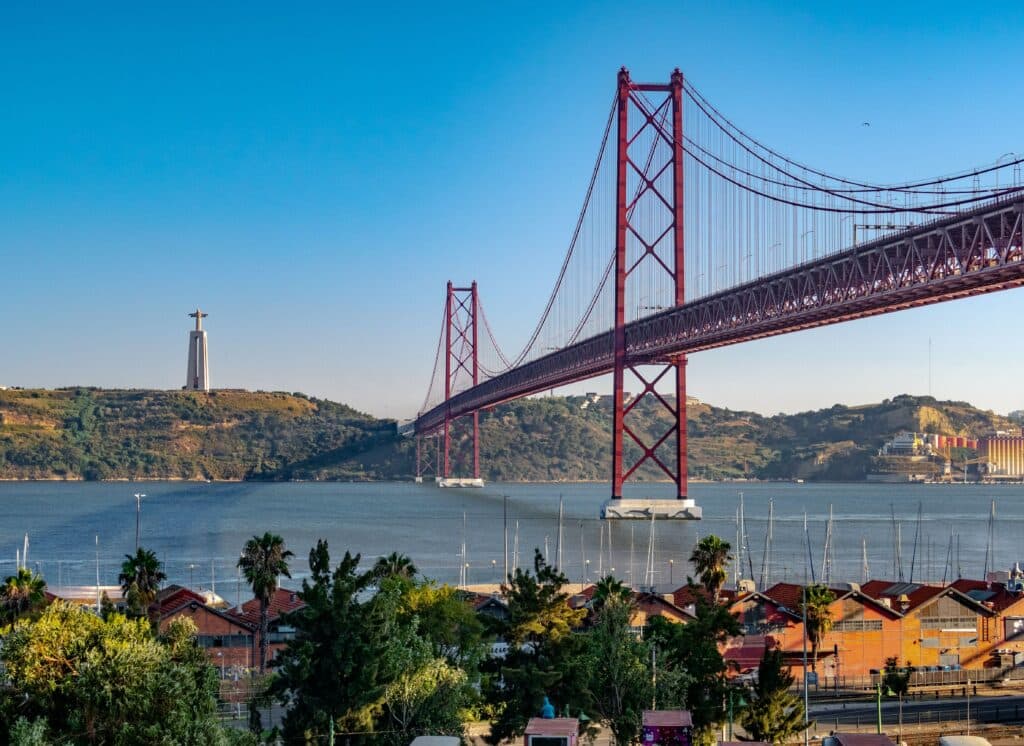 Vista da Ponte 25 de Abril, em Lisboa, Portugal, durante o dia com casas a frente em meio a arvores e ao fundo o rio e a ponte.