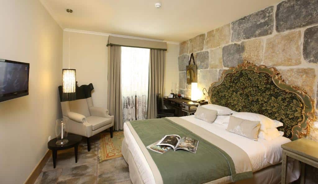 Quarto elegante do Solar do Castelo com cama grande, cabeceira trabalhada, poltrona, secretária e parede de pedra.
