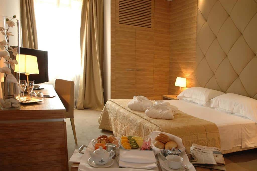 quarto do Garden Area Roma Eur, um dos hotéis baratos em Roma, com pequeno-almoço farto servido no quarto sobre uma bancada com cadeira de madeira, televisão em frente à cama com pormenores em dourado claro, mesas de cabeceira e candeeiro de ambos os lados, papel de parede texturado e janela grande com cortinados