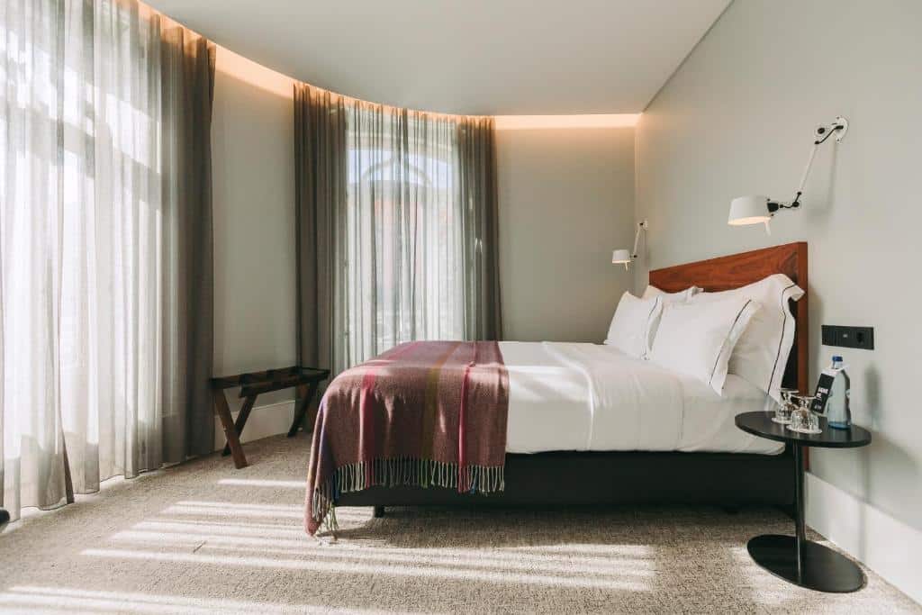 Quarto do 1908 Lisboa Hotel, com cama de casal e almofadas e janelas com cortinas em frente e à esquerda da cama