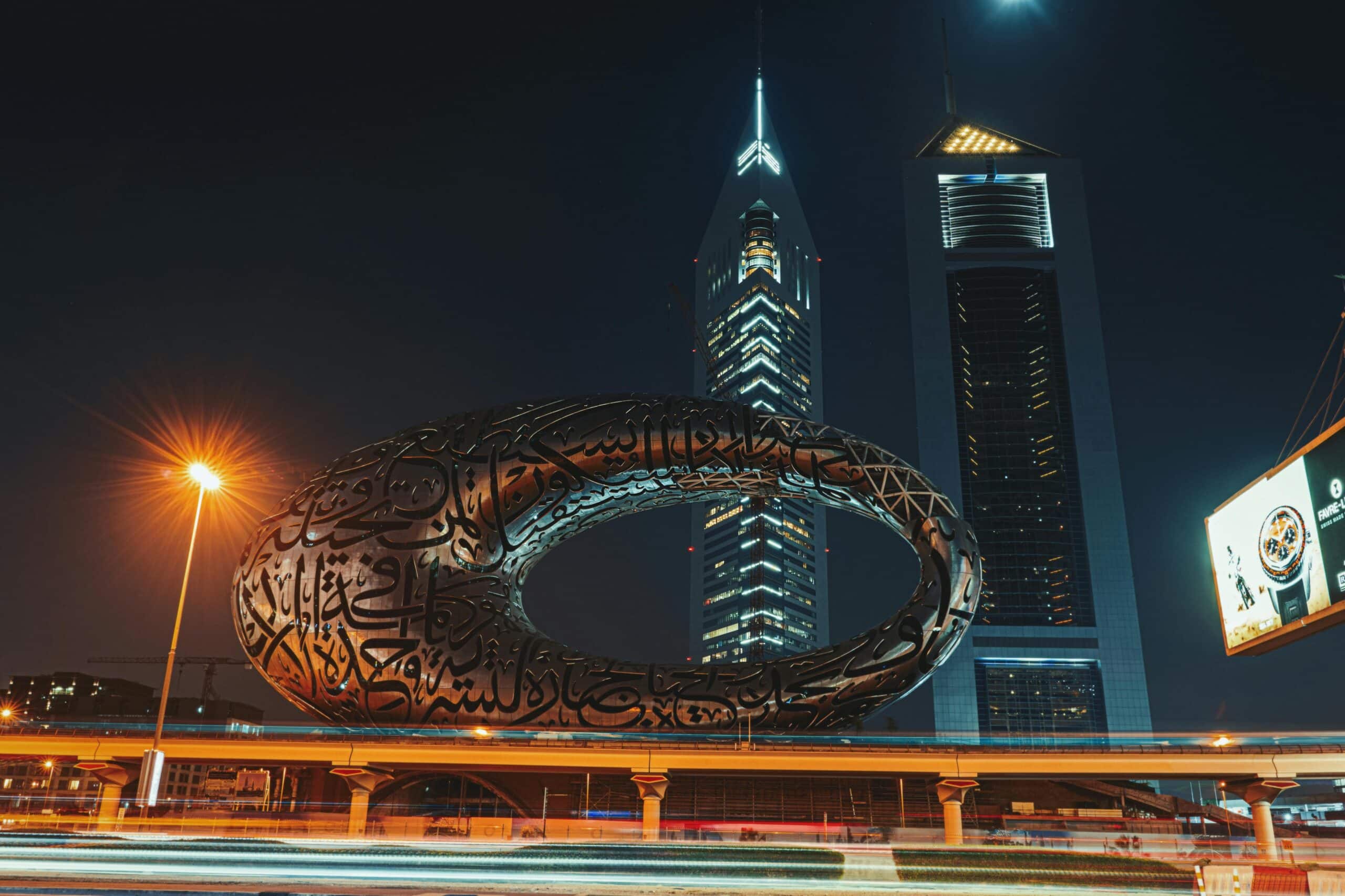 Edifício futurista com caligrafia árabe na fachada, no Museu do Futuro, com arranha-céus iluminados ao fundo.