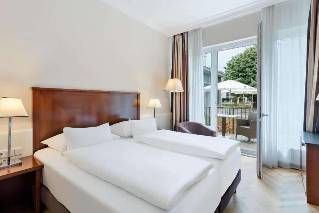 Quarto no Austria Trend Parkhotel Schönbrunn Wien, com cama de casal, dois candeeiros, uma cadeira e uma porta aberta para uma varanda com assentos ao ar livre.