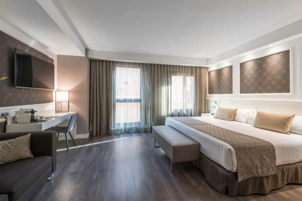 Quarto do Catalonia Magdalenes. A cama tem lençóis e almofadas bege e brancos, com candeeiros nas mesas de cabeceira, secretária com máquina de café e um cadeirão castanho ao lado.