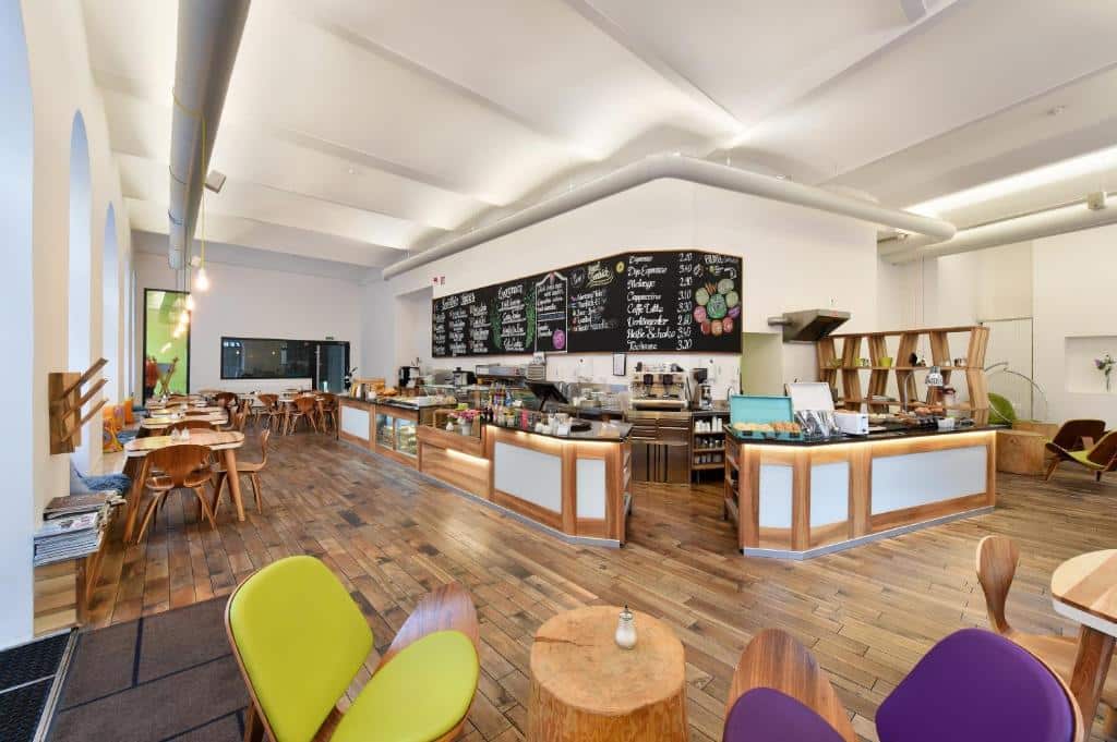 Interior de uma cafetaria moderna no Hotel Mocca, com chão de madeira, cadeiras coloridas, mesas de madeira e uma ementa em ardósias atrás do balcão.