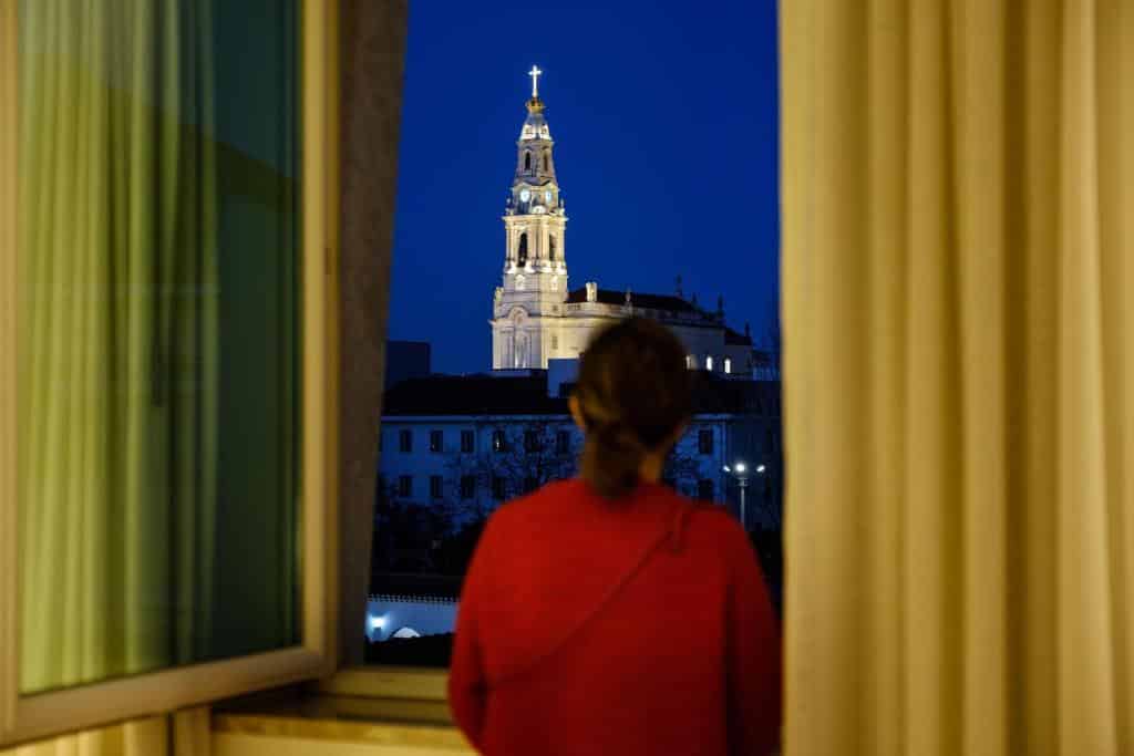Pessoa de top vermelho a olhar pela janela aberta para a torre iluminada de uma igreja com uma cruz, à noite, no Hotel Santa Maria.