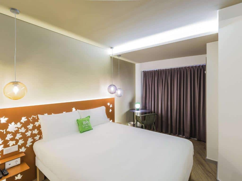 Quarto do Ibis Styles Lisboa Centro Marquês de Pombal com cama grande, decoração de parede com folhas, mesa pequena com cadeira e cortinas.