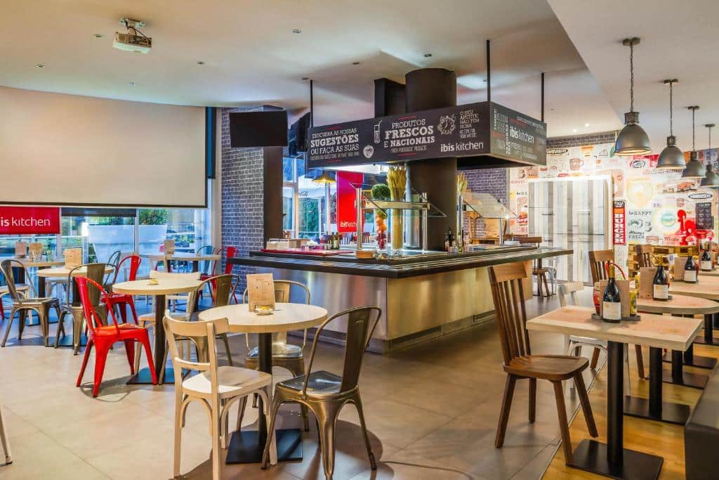 Área de refeições do Ibis Lisboa Alfragide com mesas de madeira e cadeiras coloridas e um balcão de self-service ao centro da sala