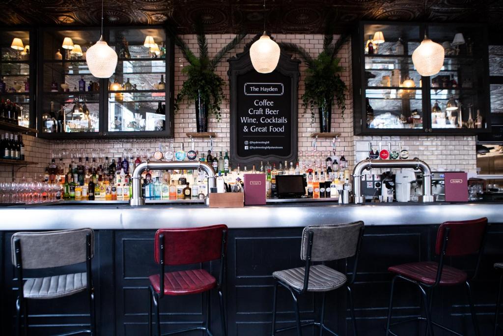 Um bar moderno com quatro cadeiras, uma variedade de garrafas, prateleiras de vidro e um quadro de menu numa parede de azulejos brancos, na propriedade do The Hayden Pub & Rooms.