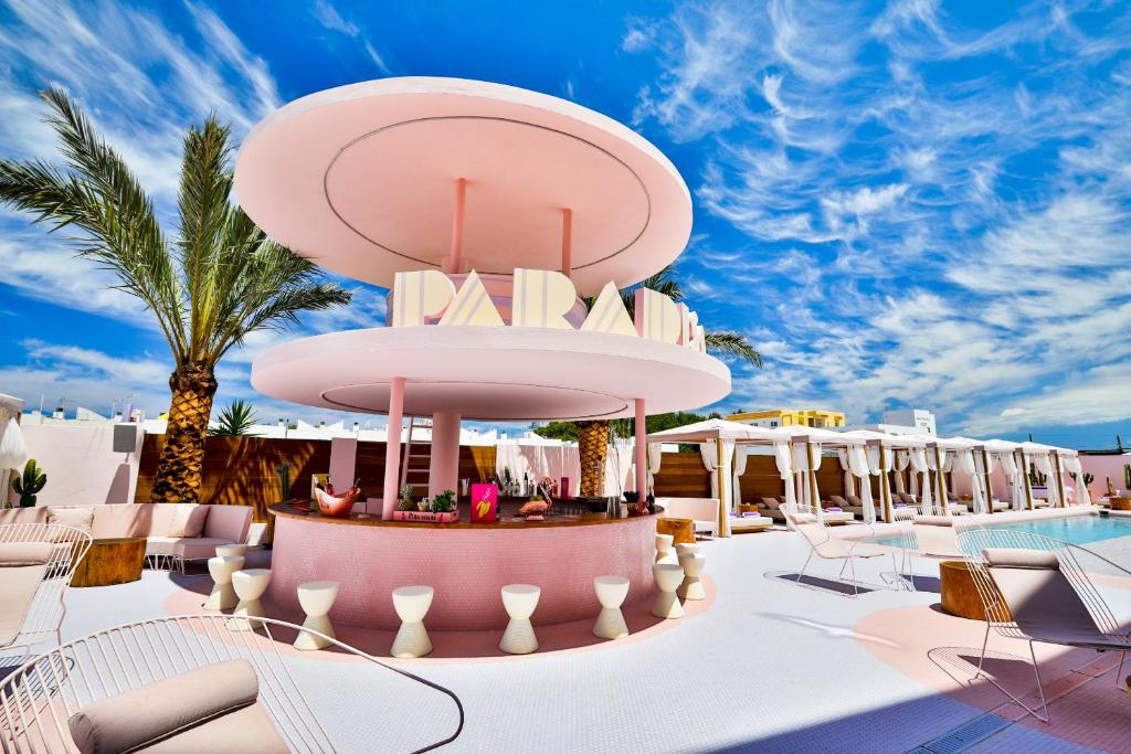 Bar circular cor-de-rosa ao ar livre no Paradiso Ibiza Art Hotel, com bancos, palmeiras e espreguiçadeiras brancas sob um céu azul.