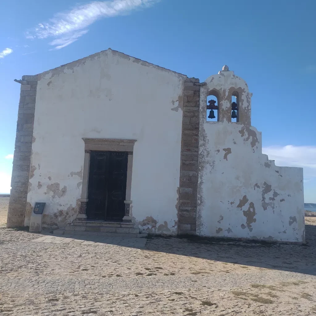 Antiga capela branca com uma torre sineira e dois sinos, sob um céu azul, num caminho de pedra.