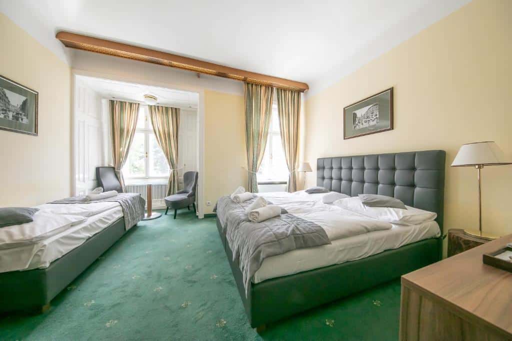 Quarto no Hotel Bergwirt Schönbrunn, com duas camas, alcatifa verde, duas janelas com cortinados, uma cadeira, candeeiros e quadros emoldurados nas paredes.