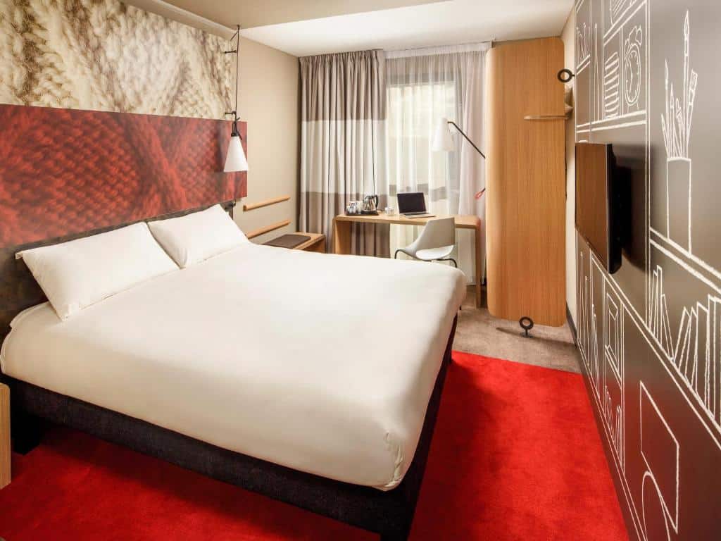 Quarto do ibis London Canning Town com cama de casal, televisão, tapete vermelho, secretária com cadeira e janela com cortinas