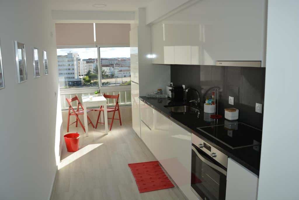 Uma cozinha moderna e estreita com armários brancos, bancadas pretas e uma pequena zona de refeições junto a uma janela com cadeiras vermelhas, no apartamento Fátima4us.