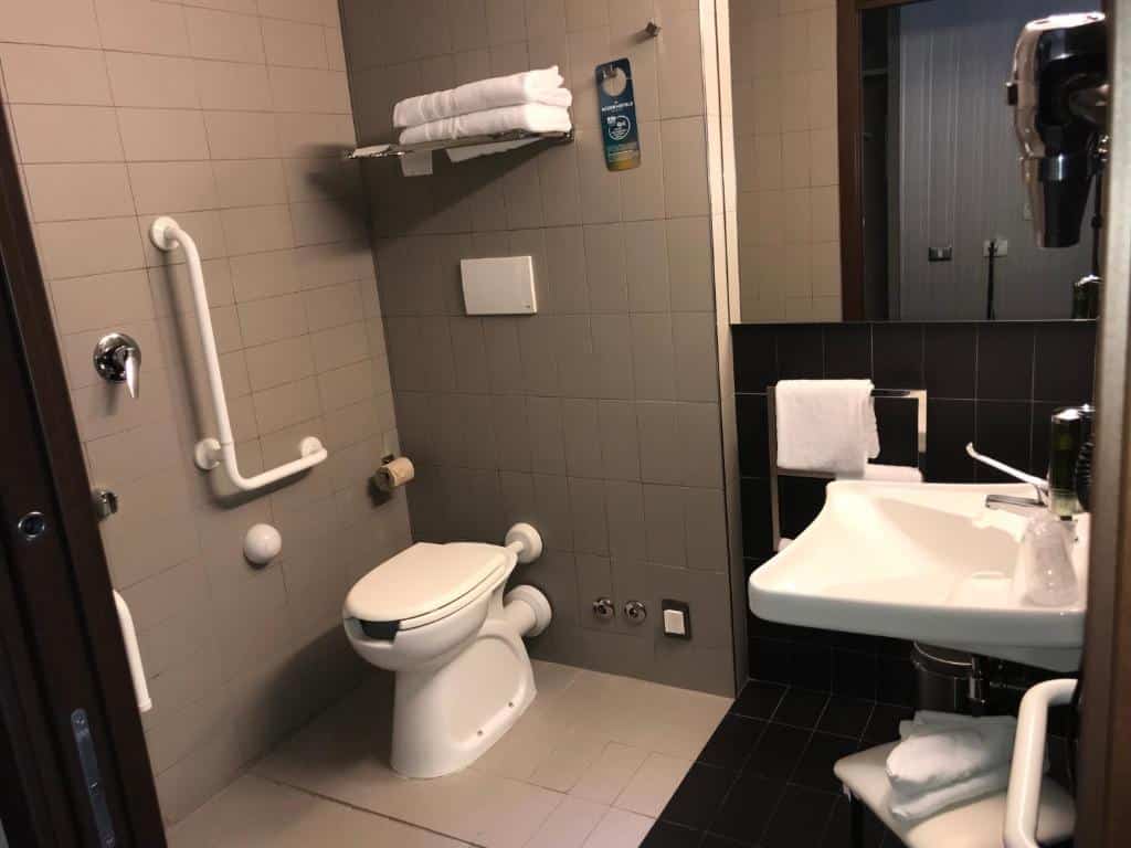 casa de banho do Ibis Styles Roma Eur, um dos hotéis baratos em Roma, com adaptações para pessoas com mobilidade reduzida: lavatório mais baixo com espaço por baixo, sanita elevada, barras de apoio e cadeira de banho num ambiente amplo