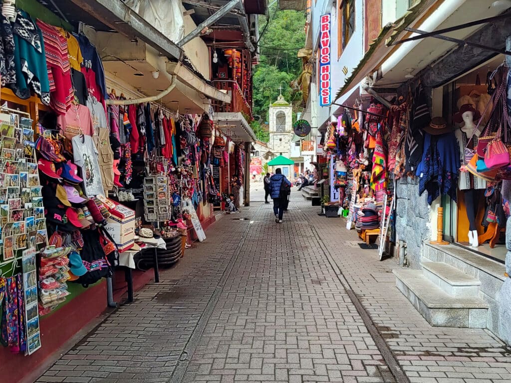 Ruas de Machupicchu Pueblo com bancas de produtos artesanais coloridos de ambos os lados e a torre da igreja ao fundo