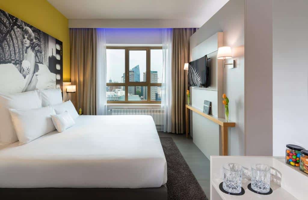 Quarto do NYX Hotel Milan by Leonardo Hotels com janela com cortinas, cama de casal, televisão e uma pequena mesa com copos e doces